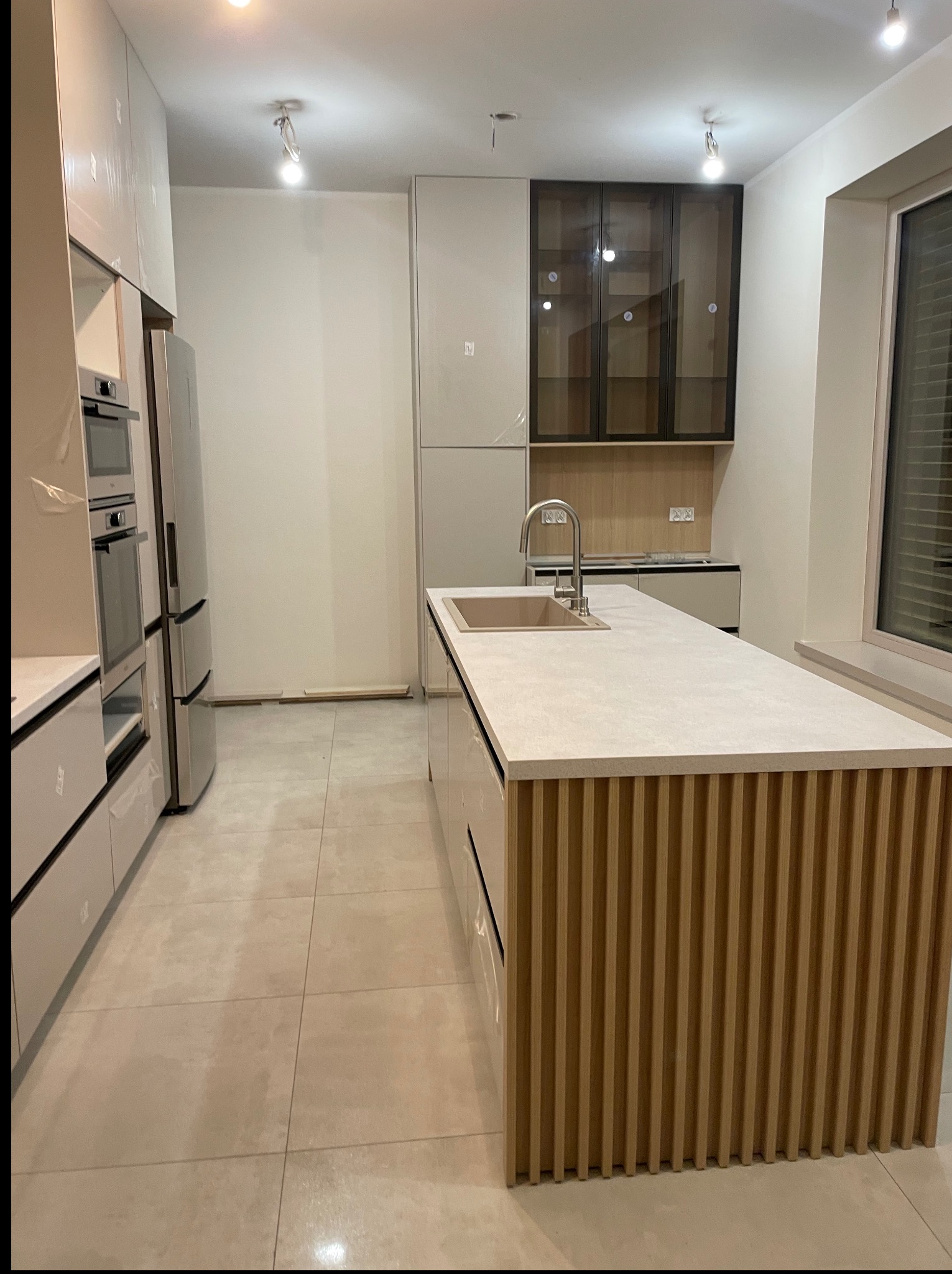 Nowoczesna kuchnia z wyspą: jasne fronty, drewniane lamele na wyspie, stalowe AGD. Minimalistyczny design, neutralna kolorystyka, duże okno z żaluzjami.