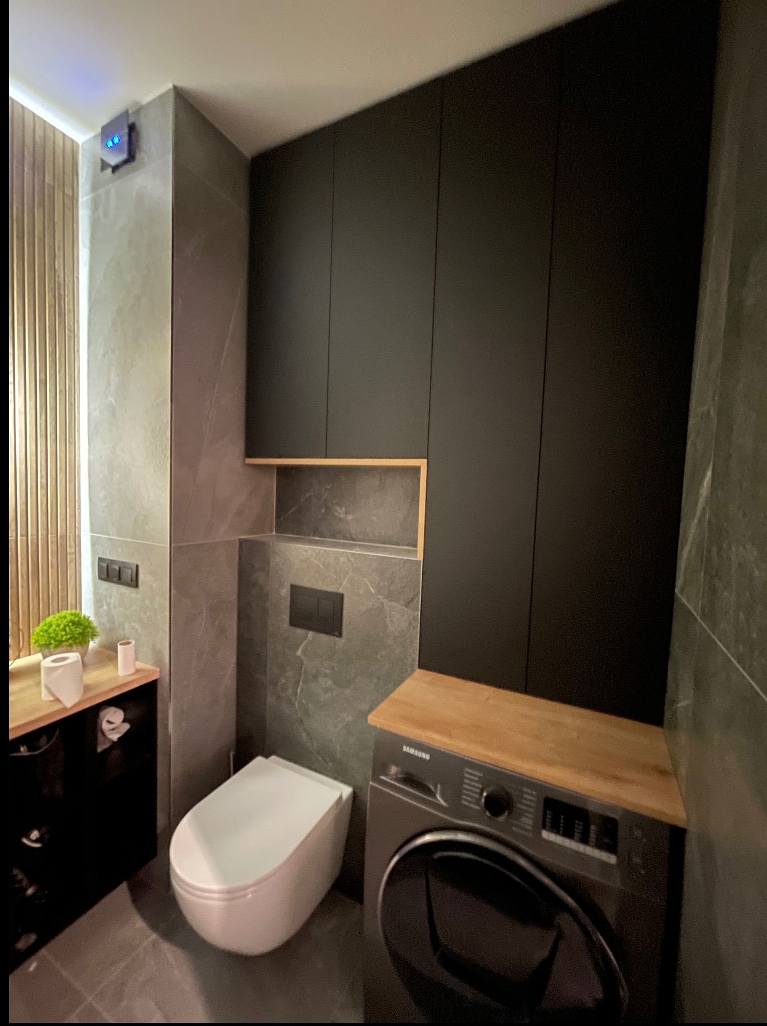 Nowoczesna łazienka z zabudową meblową: czarne szafki na wymiar, pralka pod blatem z drewna, toaleta podtynkowa i szare płytki. Minimalistyczny design.