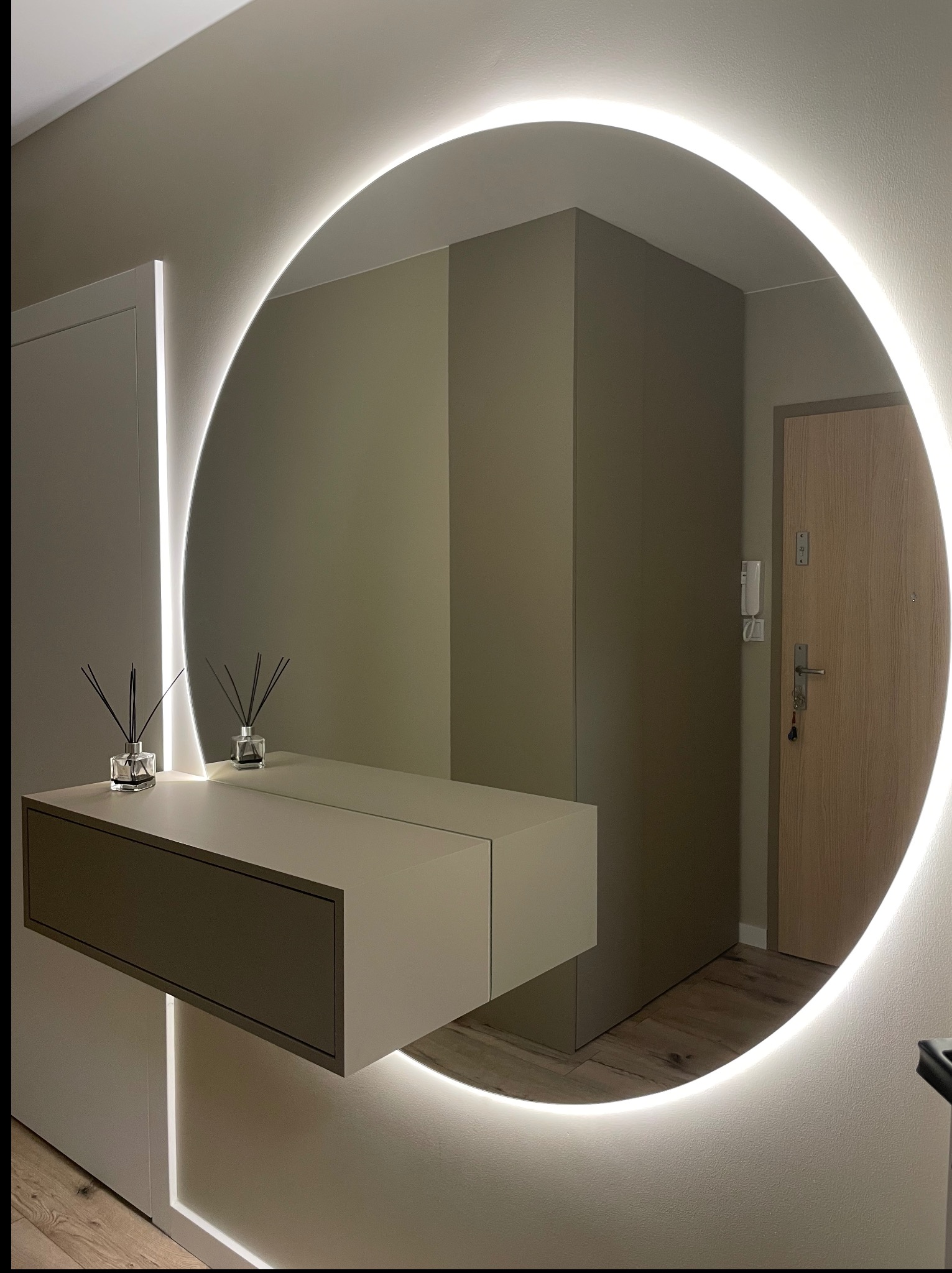 Nowoczesna szafa wnękowa z lustrem w okrągłej ramie LED i półką, minimalistyczny design, widok na drzwi wejściowe w odbiciu lustra.