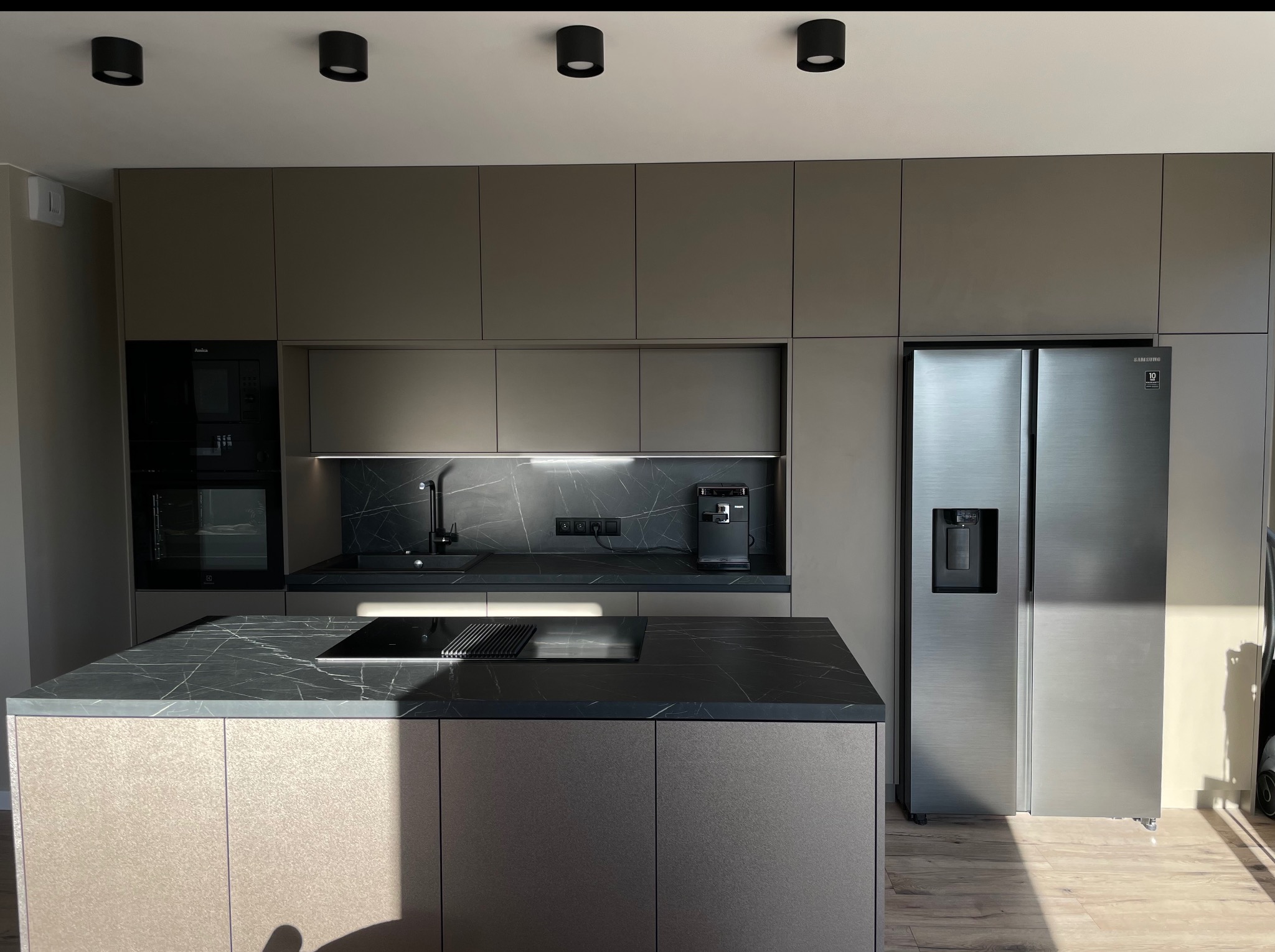 Nowoczesna kuchnia w odcieniach szarości z wyspą i blatem imitującym marmur. Na blacie płyta indukcyjna. W tle zabudowa kuchenna i duża lodówka.