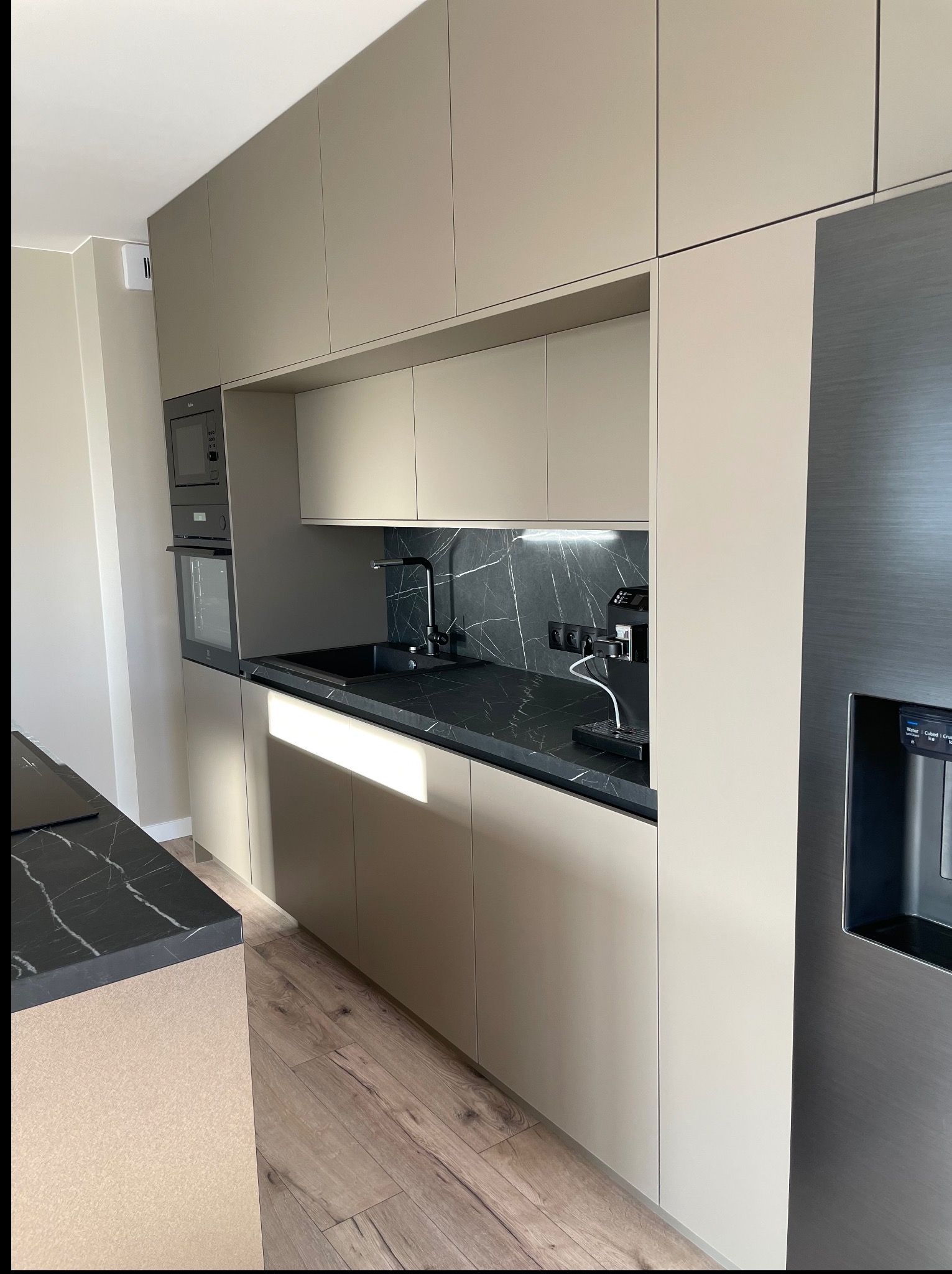 Nowoczesna kuchnia: beżowe szafki, czarny blat z marmurowym wzorem, srebrna lodówka z dystrybutorem wody. Oświetlenie punktowe nad blatem i drewniana podłoga.