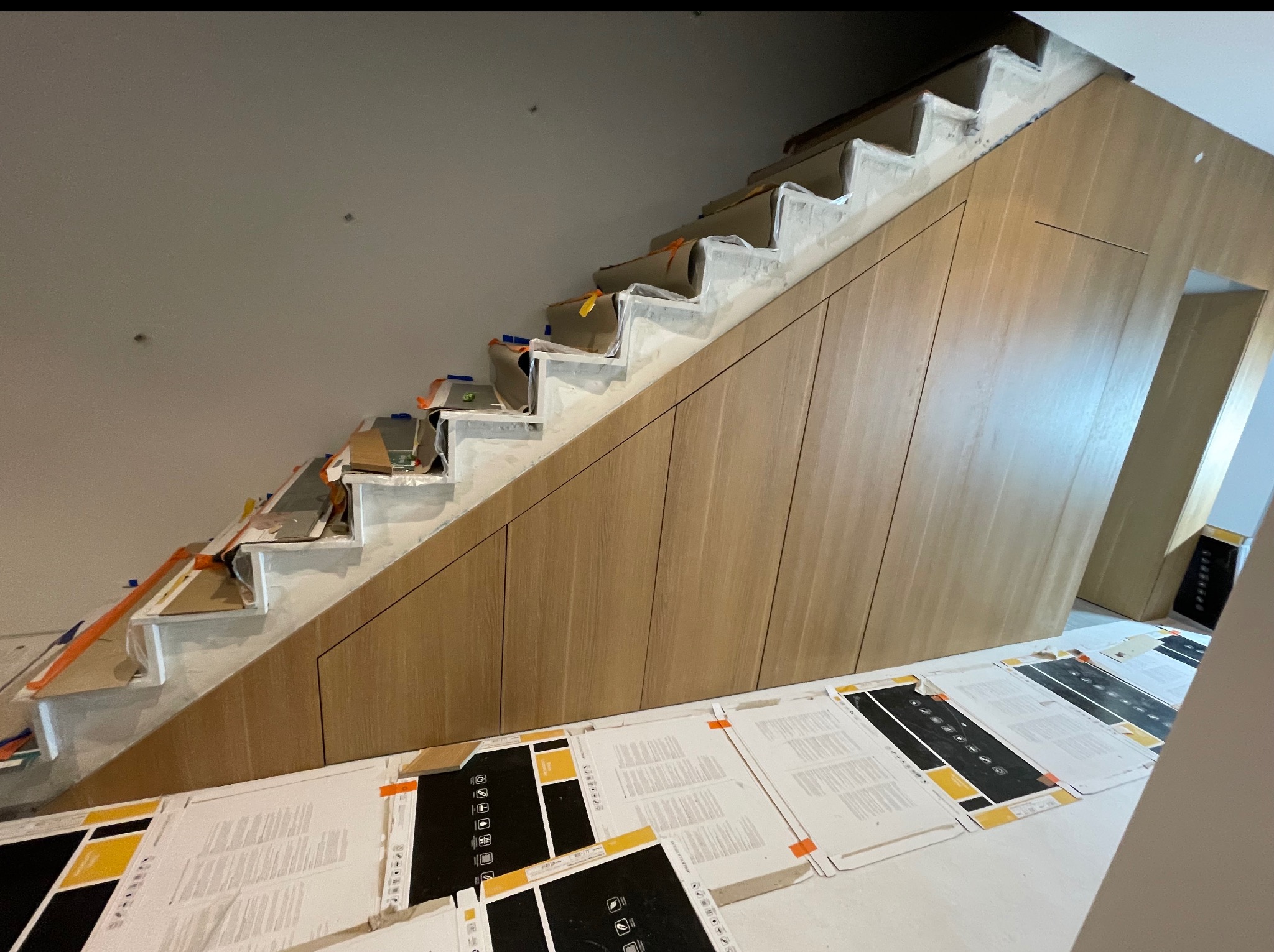 Szafa wnękowa pod schodami w trakcie montażu. Widoczne zabezpieczone schody, drewniane fronty szafy i materiały montażowe na podłodze.