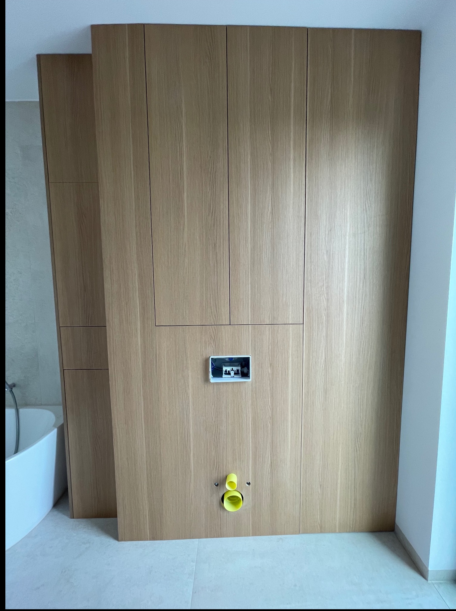 Drewniana zabudowa w łazience z ukrytym systemem spłukiwania i żółtymi elementami instalacji. Obok widoczna wanna i fragment szafki.