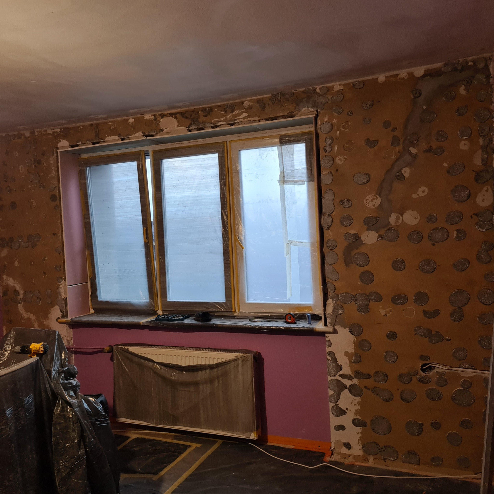 Przygotowanie ścian do szpachlowania: ściany z widocznymi punktami po kuciu, okno zabezpieczone folią, grzejnik osłonięty, narzędzia na parapecie. Etap prac remontowych.