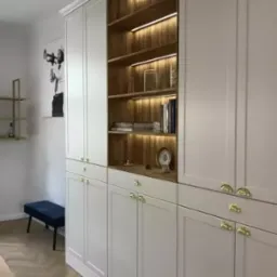 Biała zabudowa meblowa z drewnianymi półkami podświetlonymi LED, złote uchwyty, parkiet w jodełkę, minimalistyczny wystrój.