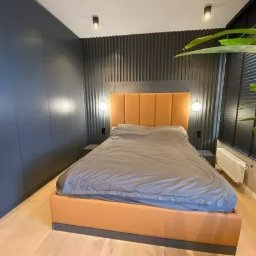 M.K.Bygg Michał Kaczmarski - Nowoczesna sypialnia z łóżkiem tapicerowanym w odcieniach beżu i szarości, na tle ściany z ciemnoszarymi lamelami i szafą w zabudowie. Minimalistyczny design, drewniana podłoga.