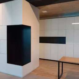 M.K.Bygg Michał Kaczmarski - Nowoczesna zabudowa meblowa z białymi szafkami i czarnym, wnękowym elementem, oświetlenie LED, minimalistyczny design, widok na ekspozycję meblową.