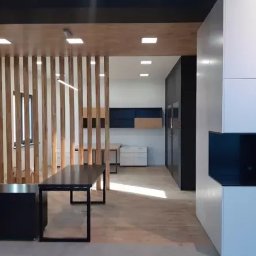 M.K.Bygg Michał Kaczmarski - Nowoczesne wnętrze z drewnianą ścianką działową i minimalistycznymi meblami. Widoczne biurko z czarnym blatem i biała zabudowa. Jasne oświetlenie.