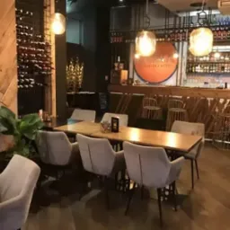 Wnętrze lokalu gastronomicznego z drewnianymi elementami: regał na wino, bar, stoły z krzesłami. Uwagę zwraca oświetlenie i rośliny doniczkowe.