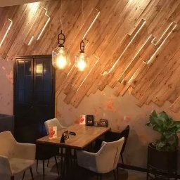 M.K.Bygg Michał Kaczmarski - Dekoracyjna ściana z desek ułożonych pod kątem, z podświetleniem LED, tworząca unikalny wzór w restauracji. Widoczne wiszące lampy i stolik z krzesłami.