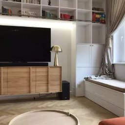 M.K.Bygg Michał Kaczmarski - Jasny salon z drewnianą komodą pod telewizorem, zabudowaną biblioteczką i siedziskiem przy oknie. Parkiet w jodełkę. Nowoczesny design, ciepłe oświetlenie.