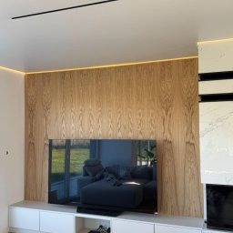 M.K.Bygg Michał Kaczmarski - Ściana TV z drewnianą okładziną, podświetleniem LED i białą szafką RTV. Widoczne połączenie drewna, bieli i czerni, tworzące nowoczesny design wnętrza.