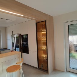 M.K.Bygg Michał Kaczmarski - Nowoczesna kuchnia z białymi frontami, drewnianym blatem i podświetlaną witryną z kieliszkami. Minimalistyczny design, jasne wnętrze, widoczny fragment krzesła barowego.