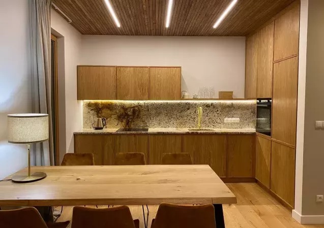 Drewniana kuchnia z podświetlanym blatem i drewnianym sufitem z oświetleniem liniowym. Stół z krzesłami na pierwszym planie. Minimalistyczny design.