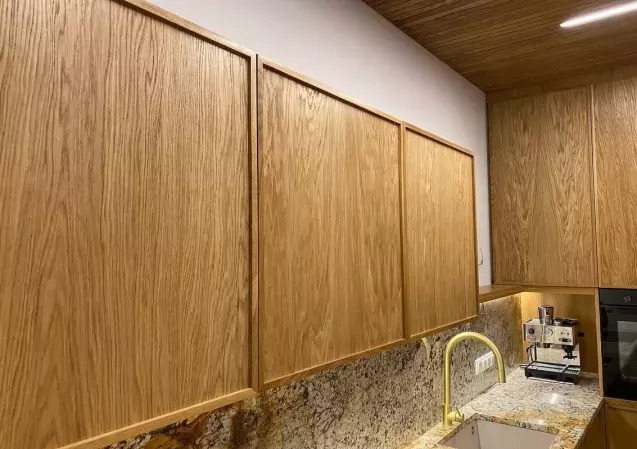 Jasne, drewniane fronty szafek kuchennych z wyraźnym usłojeniem, złota bateria przy zlewie i ekspres do kawy w tle. Ujęcie z niskiego kąta.