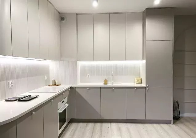 Jasnoszara kuchnia z białym blatem i podświetleniem LED. Ryflowana ściana nad blatem. Minimalistyczny design z okrągłymi uchwytami. Nowoczesne wykończenie.