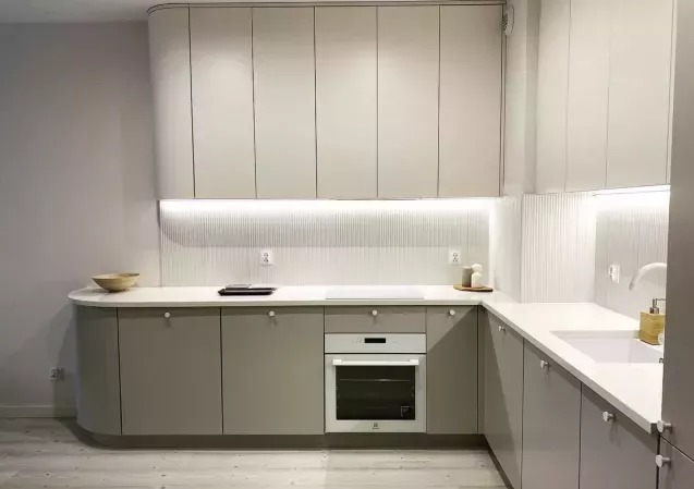 Nowoczesna kuchnia z szarymi szafkami, białym blatem i subtelnym oświetleniem LED pod szafkami wiszącymi. Minimalistyczny design z zaokrąglonymi narożnikami.