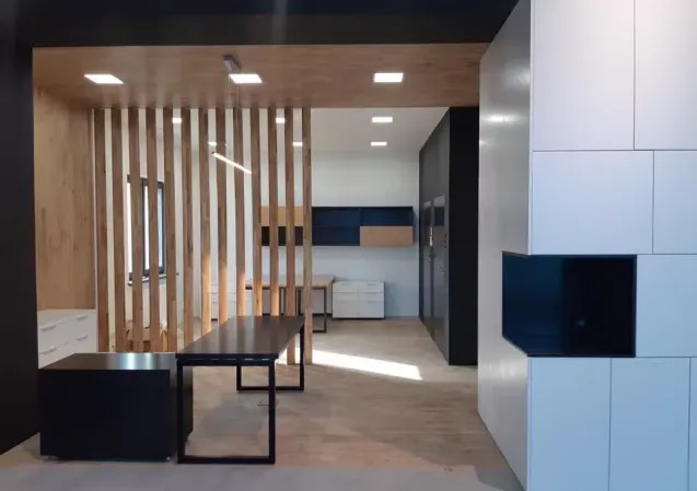 Nowoczesne wnętrze z drewnianą ścianką działową i minimalistycznymi meblami. Widoczne biurko z czarnym blatem i biała zabudowa. Jasne oświetlenie.