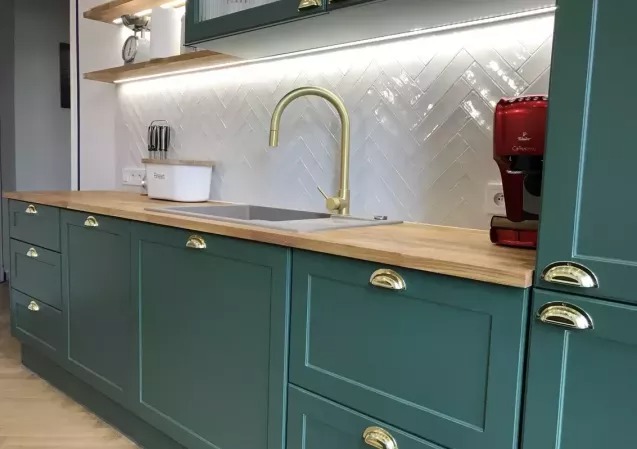Zielona kuchnia na wymiar z drewnianym blatem i złotą armaturą. Uwagę przykuwa wzór jodełki na białych płytkach ściennych i czerwony ekspres do kawy.