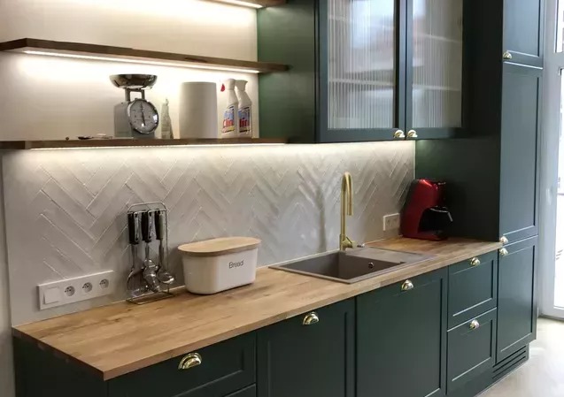 Zielona kuchnia z drewnianym blatem, złotą armaturą i frezowanymi frontami. Na półkach waga kuchenna i środki czystości. Nad blatem oświetlenie LED.