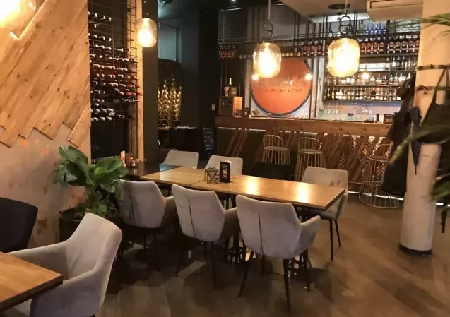 Wnętrze lokalu gastronomicznego z drewnianymi elementami: regał na wino, bar, stoły z krzesłami. Uwagę zwraca oświetlenie i rośliny doniczkowe.