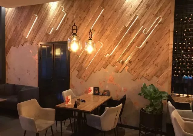 Dekoracyjna ściana z desek ułożonych pod kątem, z podświetleniem LED, tworząca unikalny wzór w restauracji. Widoczne wiszące lampy i stolik z krzesłami.