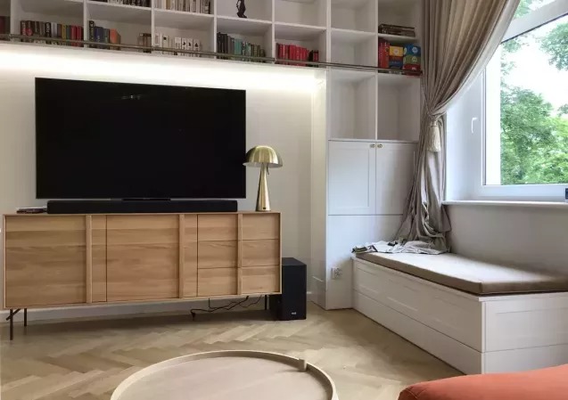 Jasny salon z drewnianą komodą pod telewizorem, zabudowaną biblioteczką i siedziskiem przy oknie. Parkiet w jodełkę. Nowoczesny design, ciepłe oświetlenie.