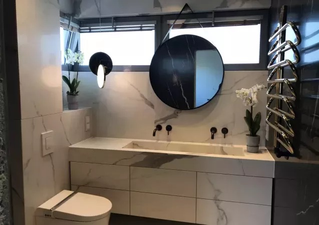 Nowoczesna łazienka z marmurową zabudową umywalki i okrągłym lustrem. Minimalistyczne czarne baterie i grzejnik w kształcie wieszaka.