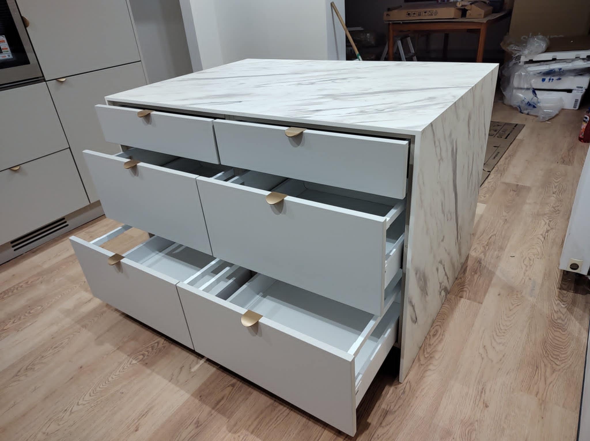 Wysuwana szuflada z szarymi frontami i marmurowym blatem, widoczna wnęka i wnętrze szuflad, złote uchwyty, na drewnianej podłodze.