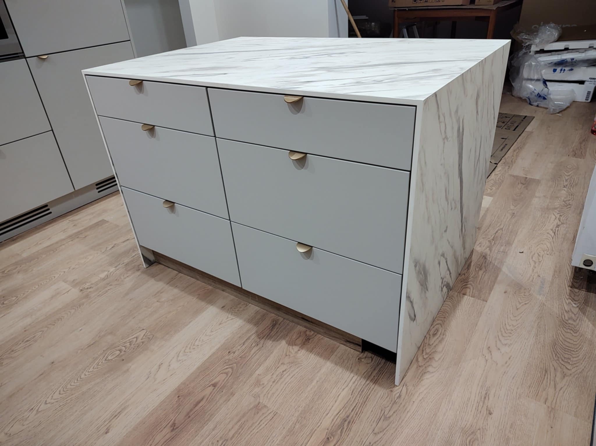 Szara wyspa kuchenna z marmurowym blatem i złotymi uchwytami na tle podłogi imitującej drewno i zabudowy kuchennej w podobnej kolorystyce.