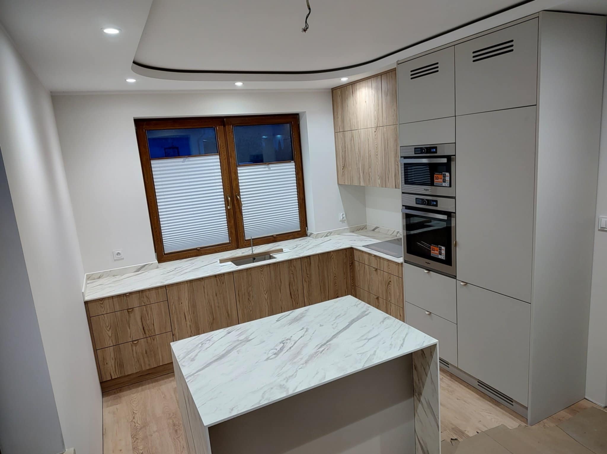 Jasna kuchnia z drewnianymi i szarymi szafkami, marmurowym blatem i wyspą. Okno z żaluzjami w tle. Nowoczesny design.