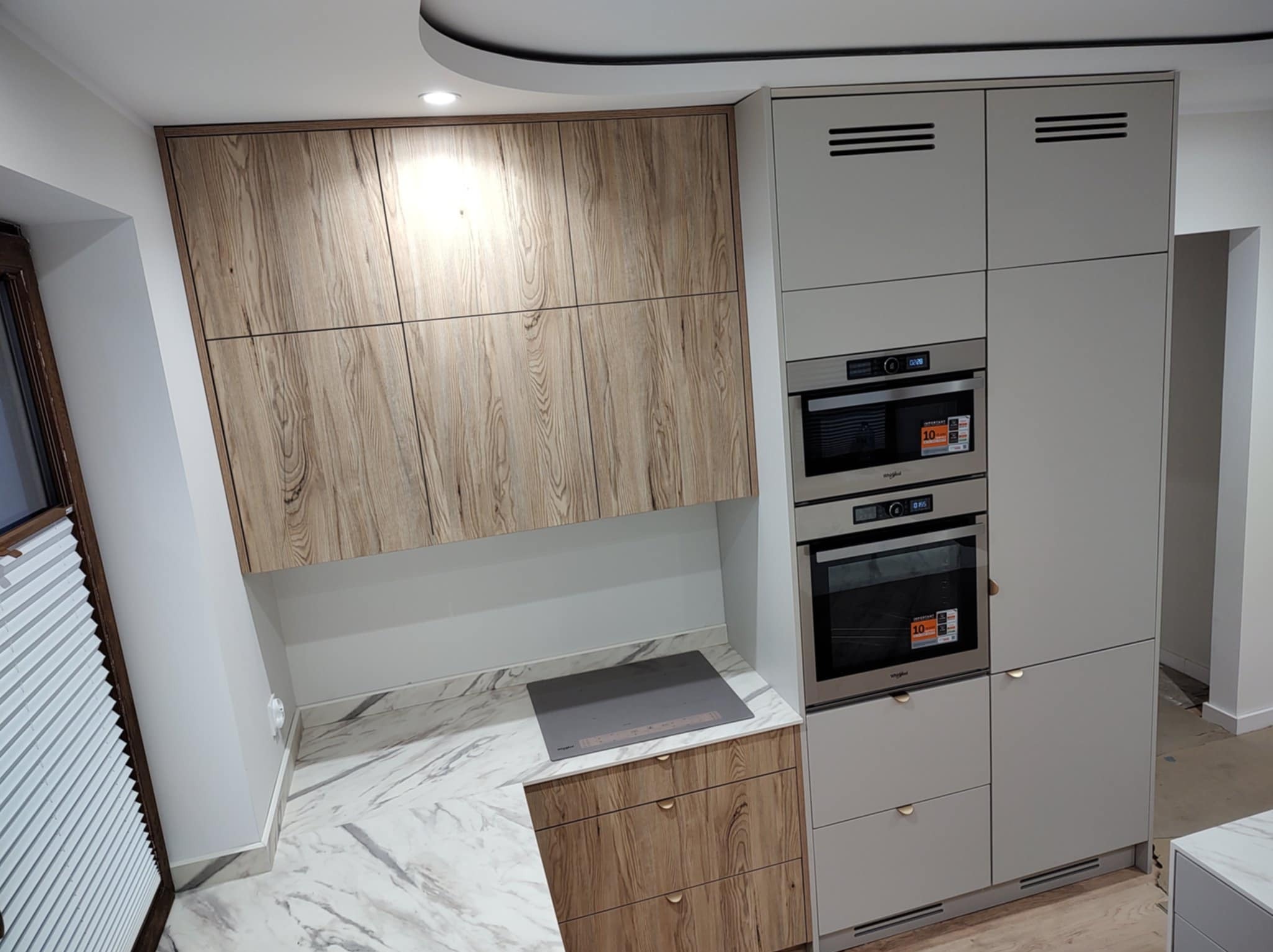 Nowoczesna kuchnia z zabudową: szafki drewnopodobne i szare, piekarnik, płyta indukcyjna na marmurowym blacie. Minimalistyczny design, widok z boku.