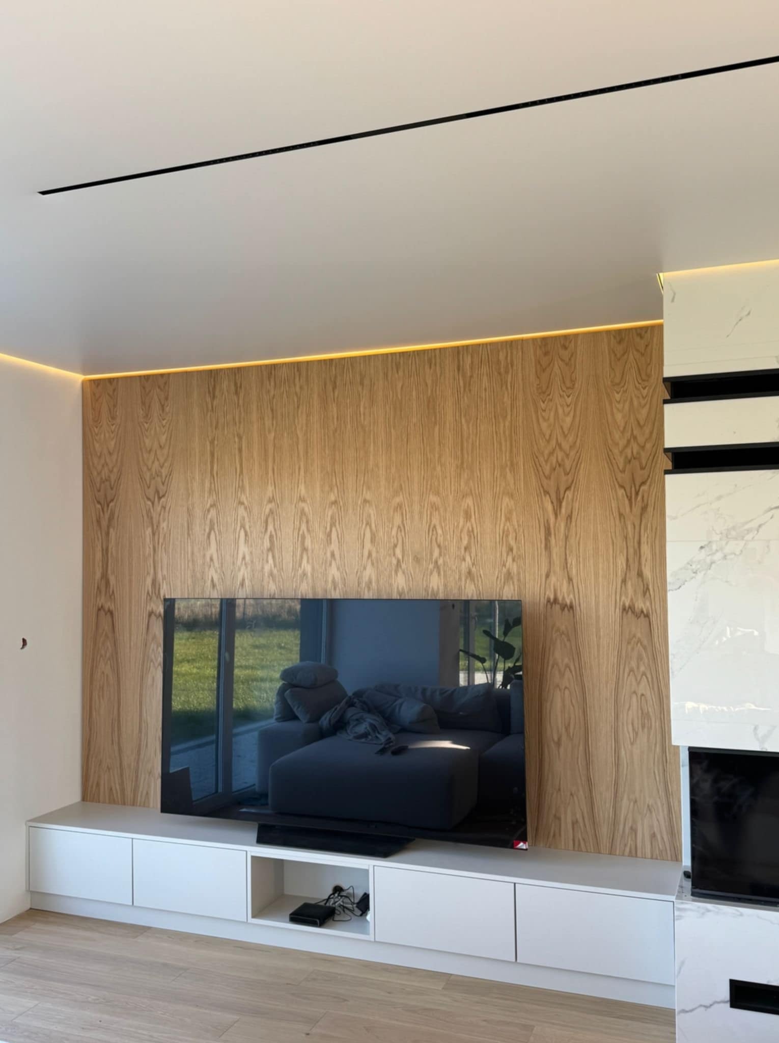 Ściana TV z drewnianą okładziną, podświetleniem LED i białą szafką RTV. Widoczne połączenie drewna, bieli i czerni, tworzące nowoczesny design wnętrza.