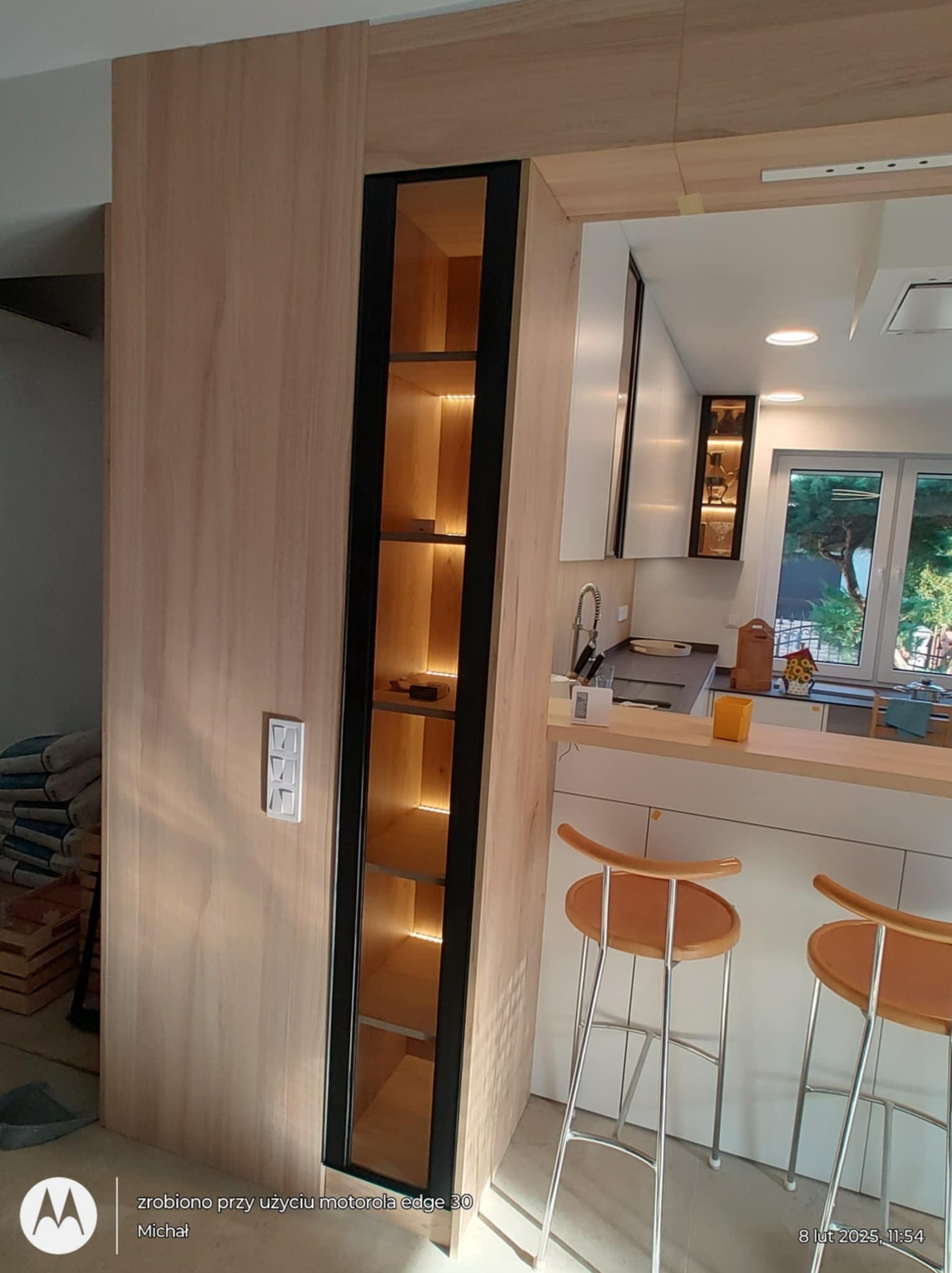 Nowoczesna kuchnia z drewnianą zabudową i czarną witryną z oświetleniem LED. Widoczne krzesła barowe i fragment blatu. Minimalistyczny design.