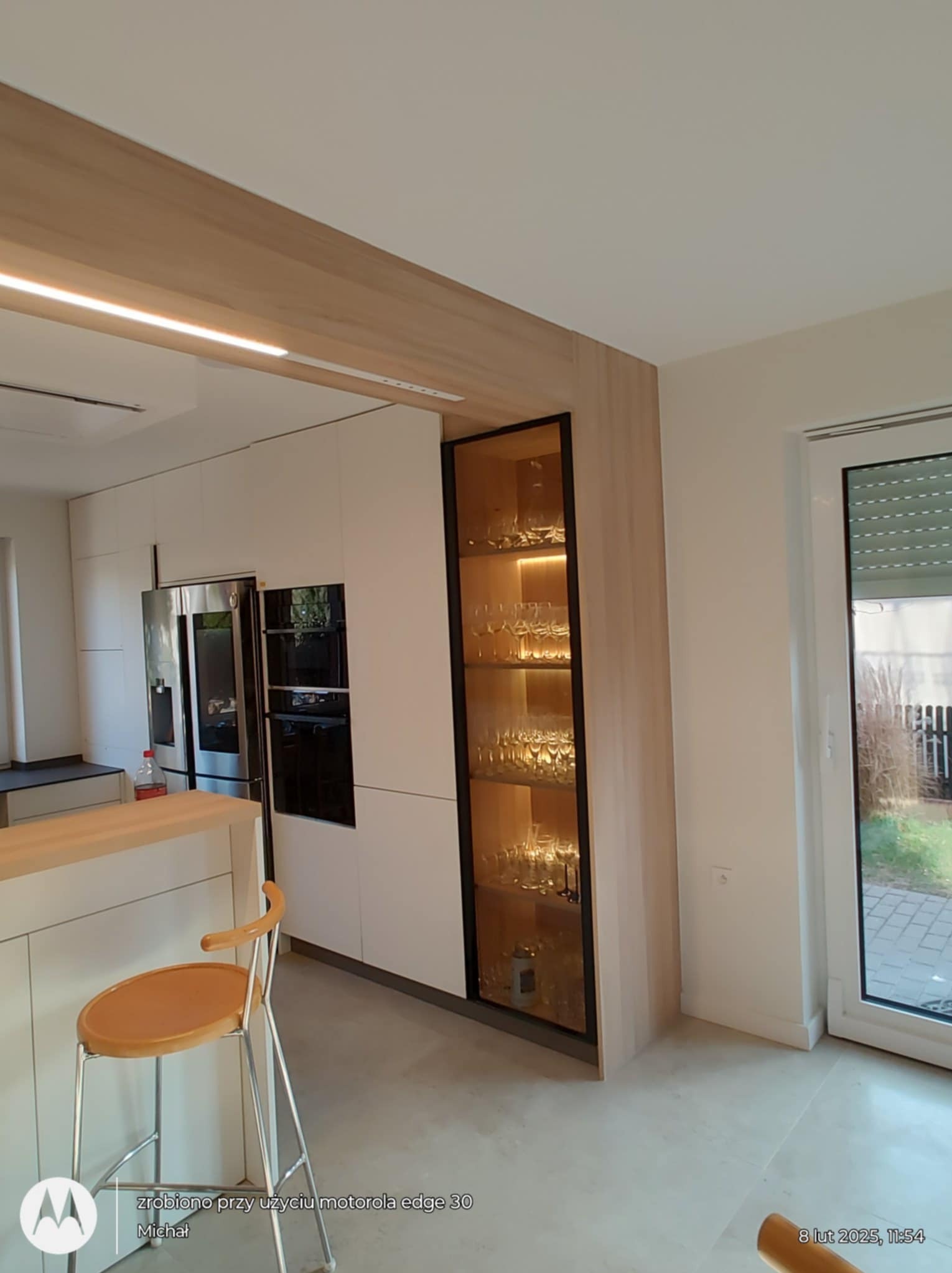 Nowoczesna kuchnia z białymi frontami, drewnianym blatem i podświetlaną witryną z kieliszkami. Minimalistyczny design, jasne wnętrze, widoczny fragment krzesła barowego.