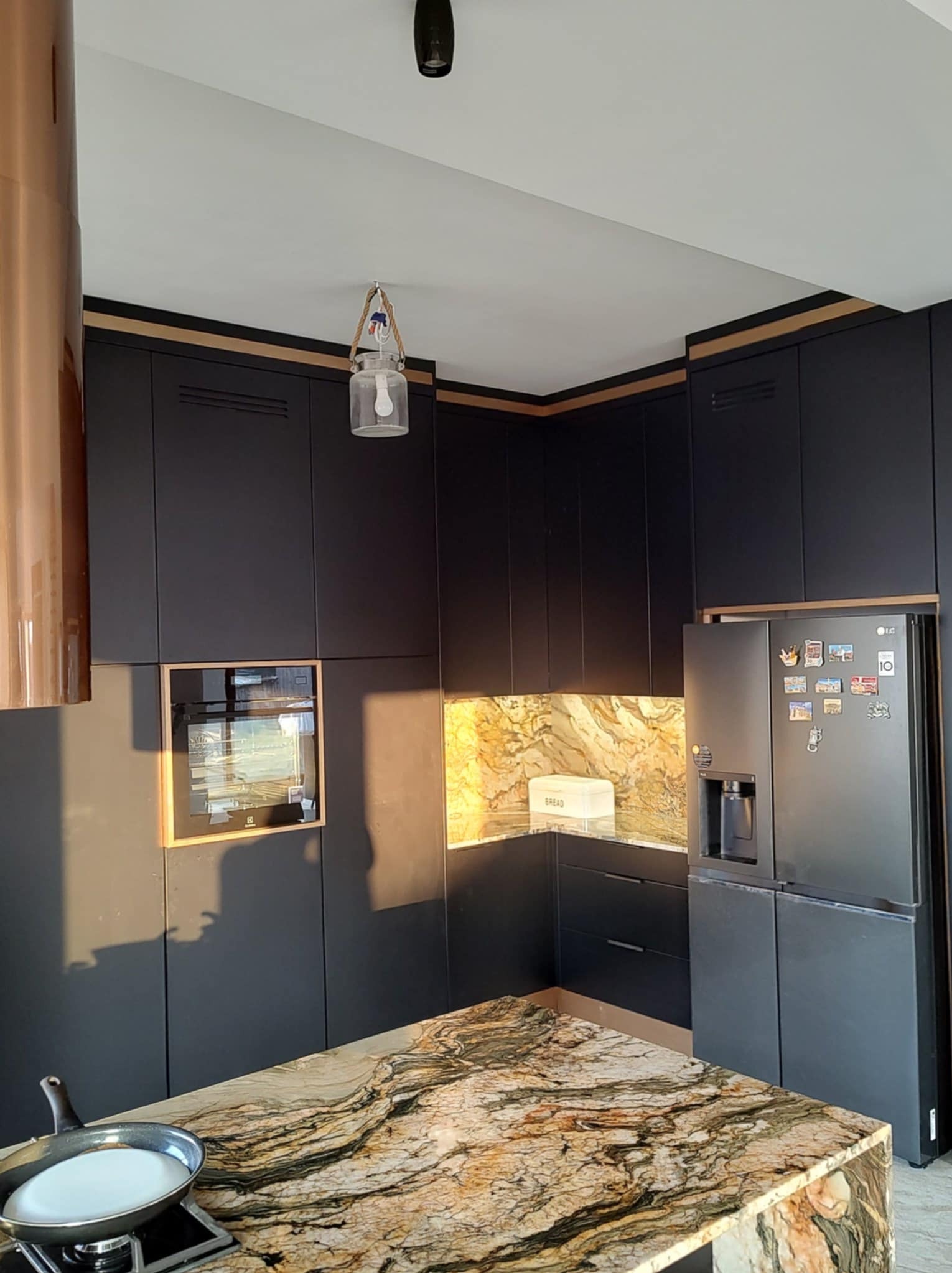 Nowoczesna kuchnia z czarnymi szafkami, marmurowym blatem i złotymi akcentami. Widoczny piekarnik, lodówka z magnesami i nietypowa lampa wisząca.