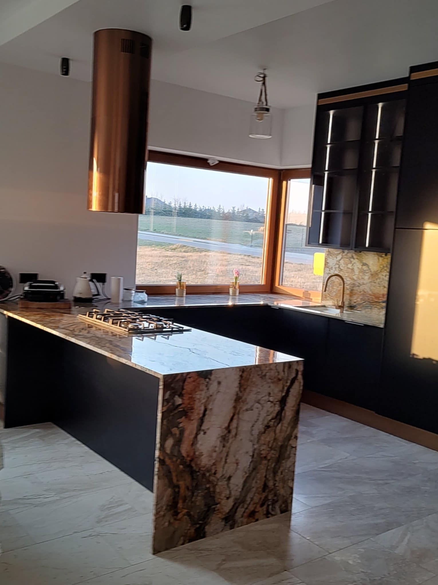 Nowoczesna kuchnia z wyspą wykończoną marmurem, okapem w kolorze miedzi i czarnymi szafkami. Duże okno z widokiem na zewnątrz doświetla przestrzeń.