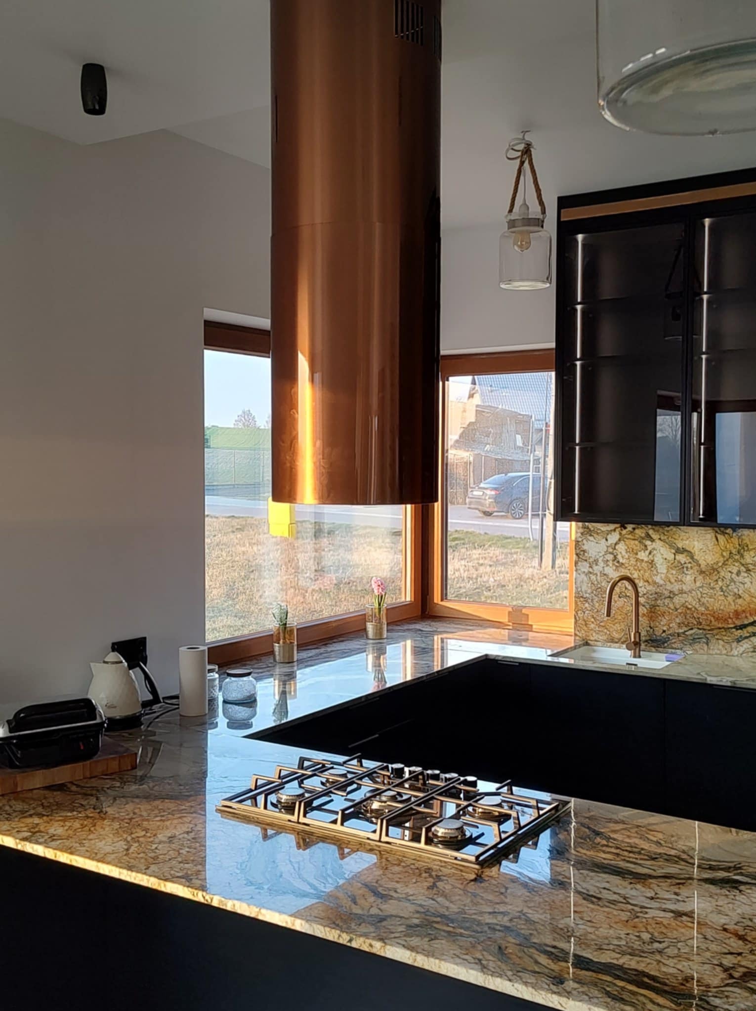 Nowoczesna kuchnia z granitowym blatem, miedzianym okapem cylindrycznym i czarnymi szafkami. Widok na zewnątrz przez okno. Elementy oświetlenia dekoracyjnego.