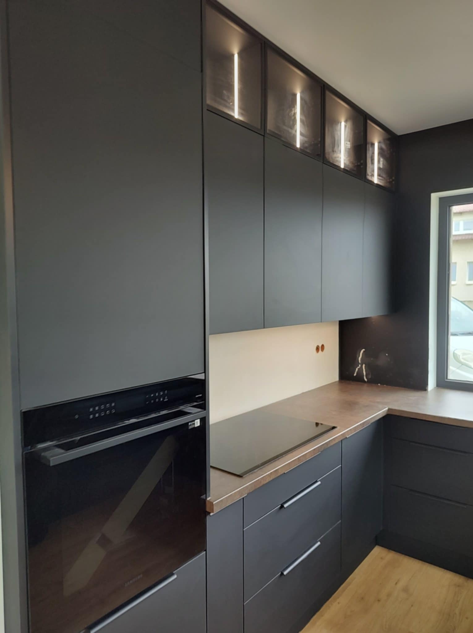 Nowoczesna, czarna kuchnia z oświetleniem LED w górnych szafkach i piekarnikiem w zabudowie. Minimalistyczny design, brązowy blat i drewniana podłoga.