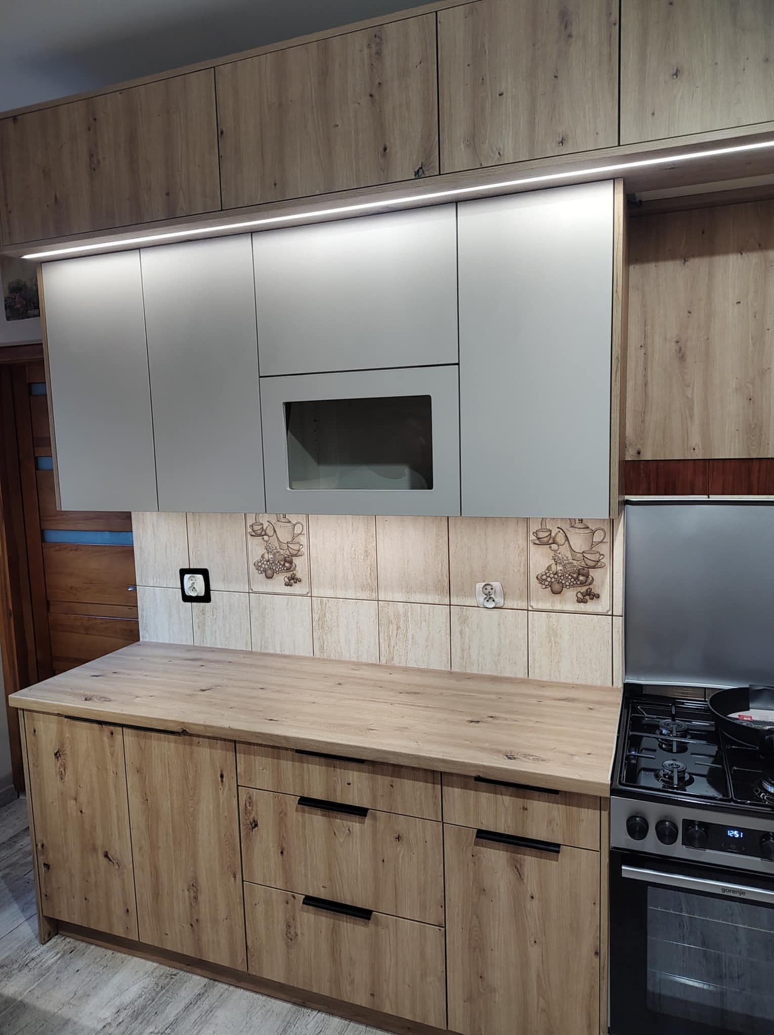 Jasna kuchnia z drewnianymi szafkami dolnymi i szarymi górnymi, podświetlona listwą LED. Blat drewniany, nad nim płytki z motywem kawy, obok kuchenka.