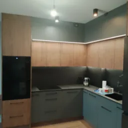 Nowoczesna kuchnia z drewnianymi i szarymi frontami, oświetleniem LED podszafkowym. Uwagę zwraca minimalistyczny design i eleganckie wykończenie. Widoczny fragment blatu i zlewu.