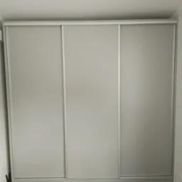 Minimalistyczna szafa wnękowa z trzema przesuwanymi drzwiami w odcieniach szarości, dopasowana do wnętrza z szarą podłogą. Widoczne narzędzia po lewej stronie.