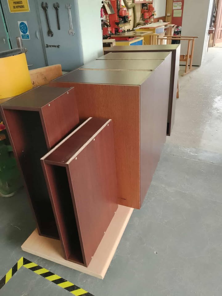 Częściowo złożone, brązowe korpusy szafek na drewnianej podstawie w warsztacie stolarskim. W tle narzędzia i maszyny stolarskie.