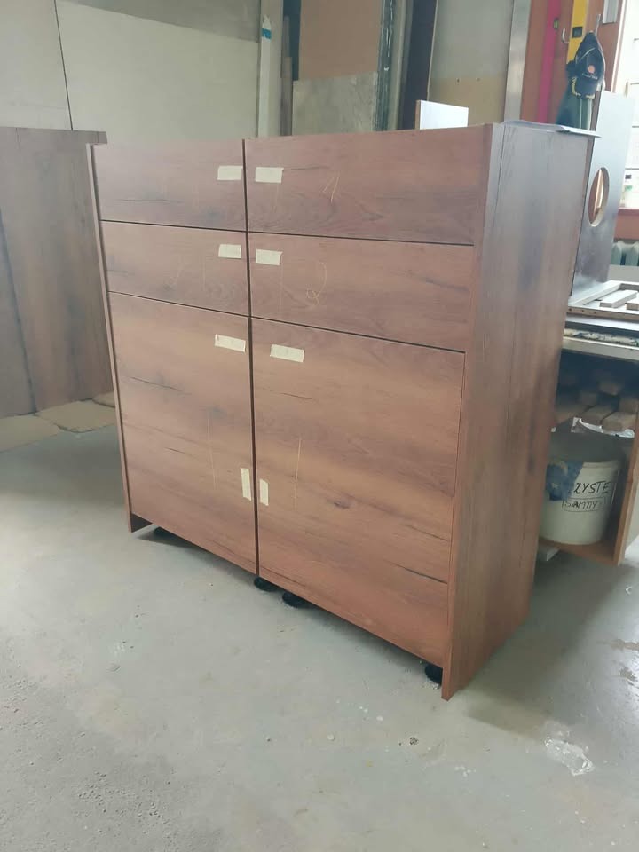 Bliższe ujęcie brązowej szafki z szufladami i drzwiczkami, z widocznymi taśmami montażowymi, stojącej w warsztacie na betonowej podłodze.