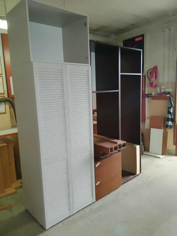 Biała szafa z lamelowymi drzwiami i otwartą nadstawką, obok elementy szafy z ciemnego drewna w warsztacie, widoczny sprzęt ppoż.