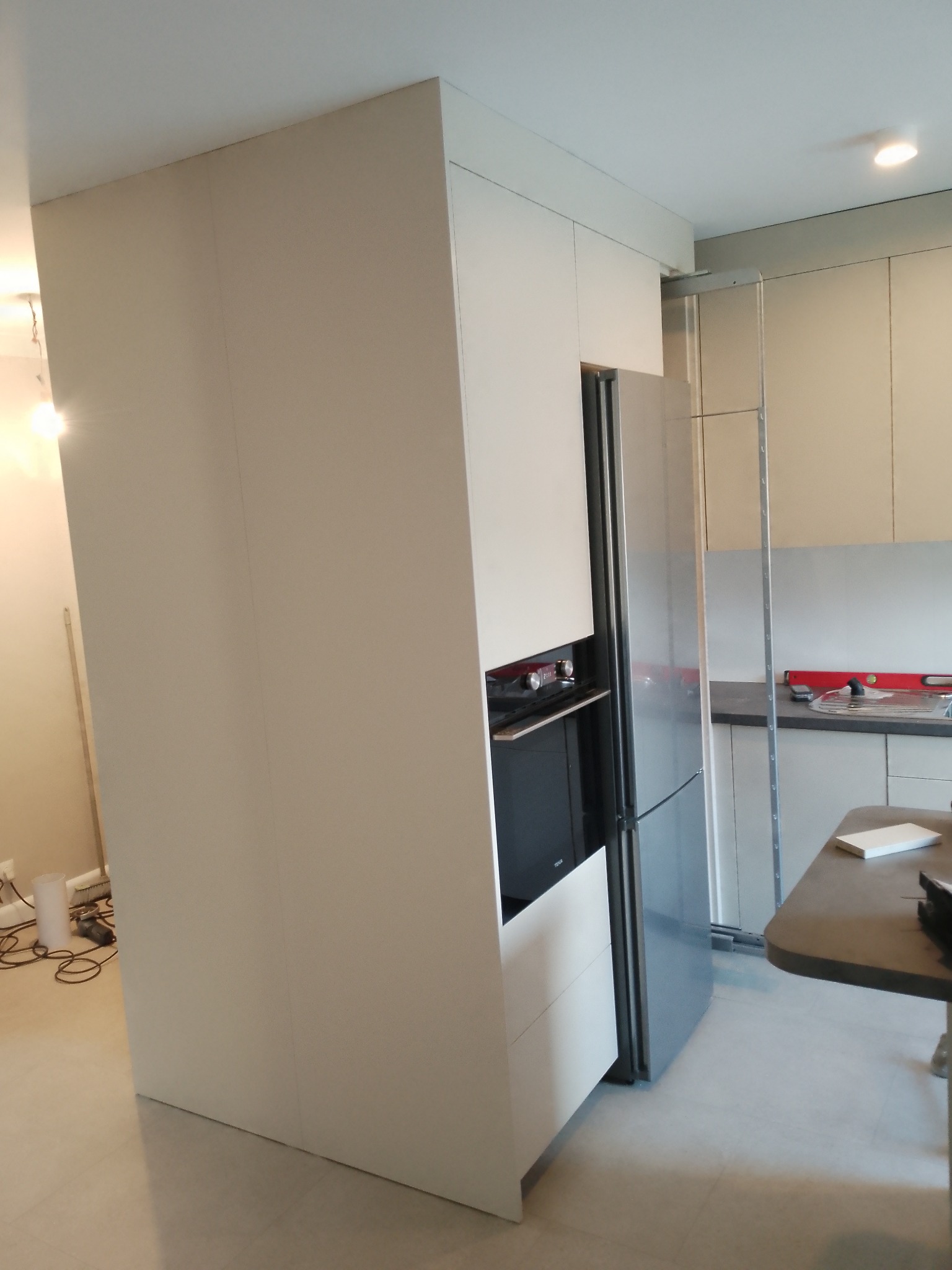 Biała zabudowa kuchenna z piekarnikiem i lodówką w nowoczesnym wnętrzu. Widoczne elementy montażowe i narzędzia.