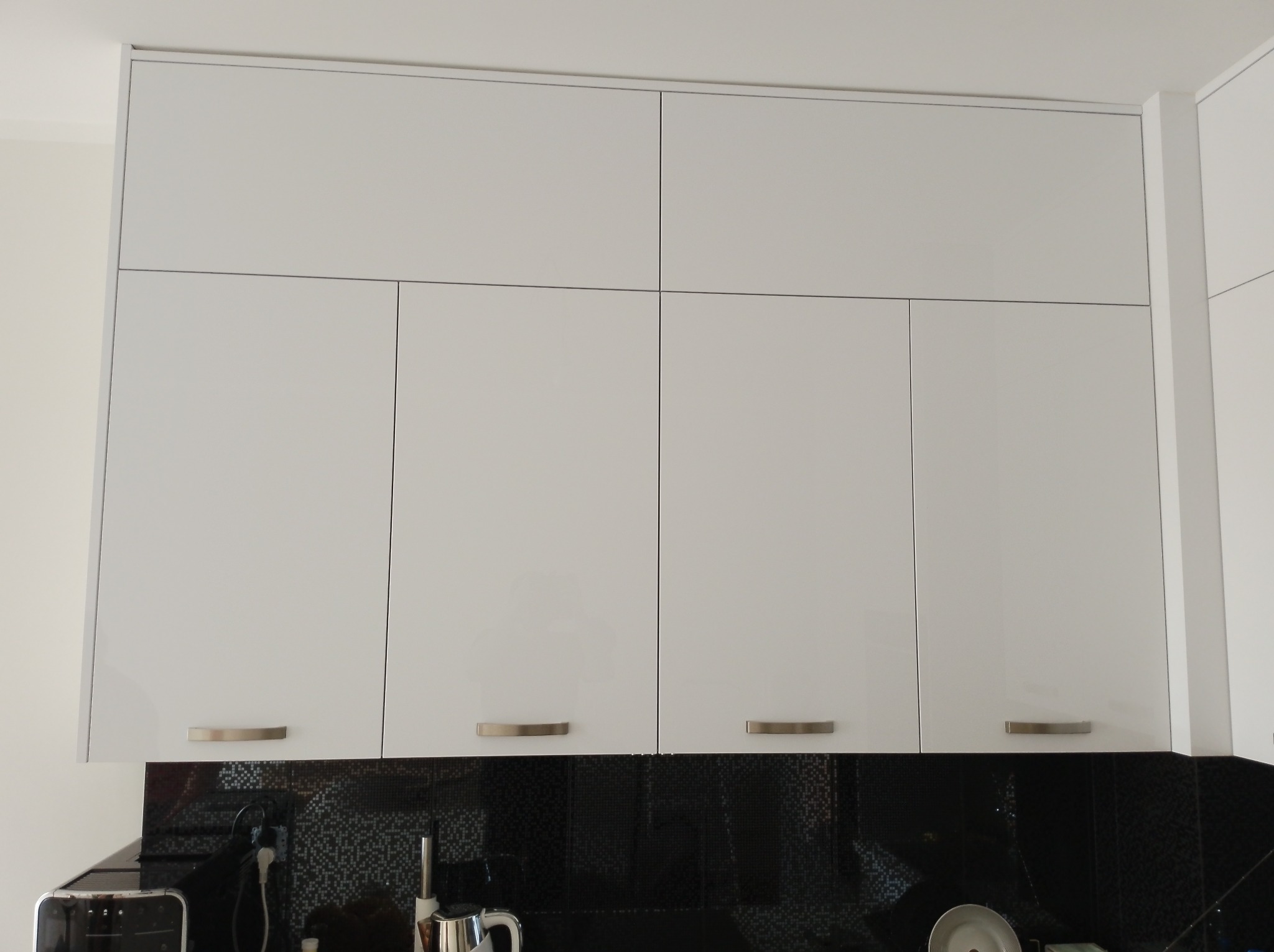 Białe, lakierowane szafki kuchenne wiszące z subtelnymi, poziomymi uchwytami na tle czarnej mozaiki. Nowoczesny design, symetryczny układ frontów.