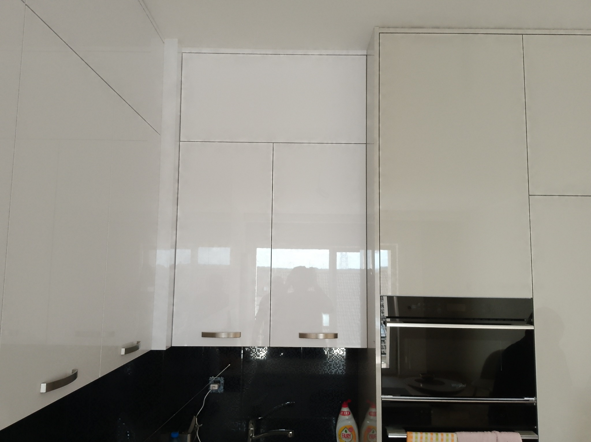 Błyszcząca, biała zabudowa kuchenna z górnymi szafkami i wbudowanym piekarnikiem na tle czarnej ściany z mozaiką. Widoczne srebrne uchwyty szafek.