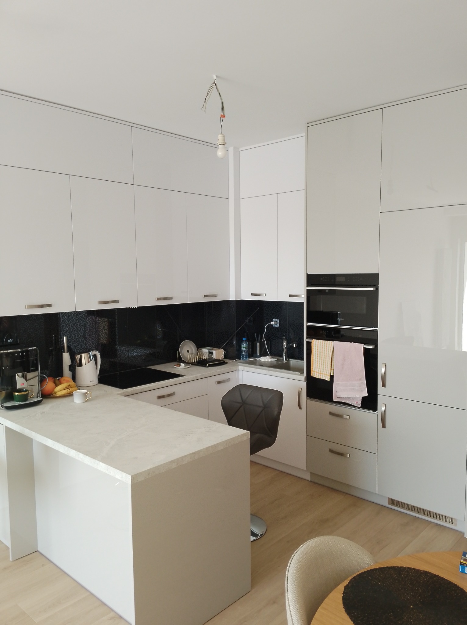 Biała, nowoczesna kuchnia na wymiar z czarnym panelem nad blatem. Widoczny barek, sprzęt AGD i wiszące przewody od oświetlenia.