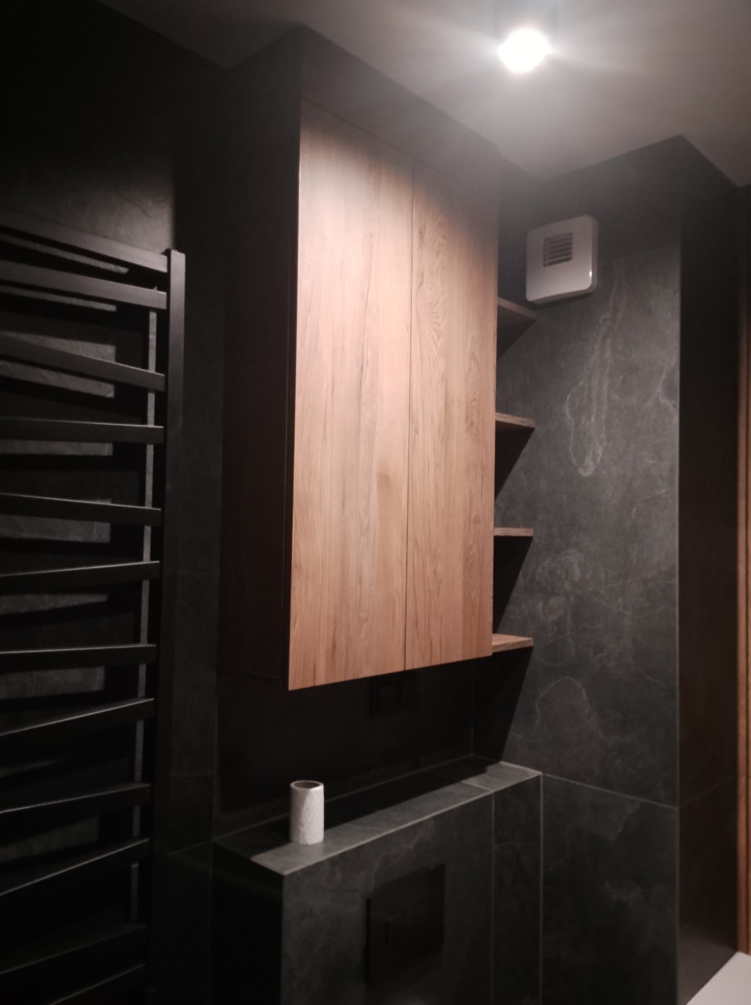 Nowoczesna łazienka z drewnianą szafką wiszącą, czarnym grzejnikiem drabinkowym i płytkami imitującymi marmur. Minimalistyczny design z akcentem na naturalne materiały.